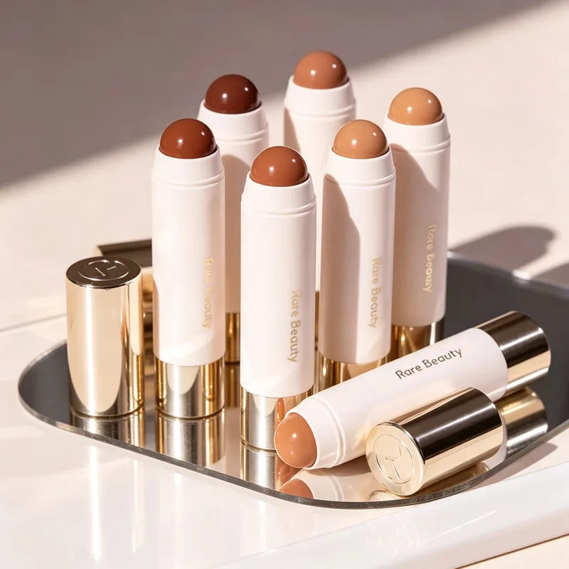 Stick Contouring nacré & Illuminateur – Contrôle Sébum by Rare Beauty in various shades displayed on a vanity.